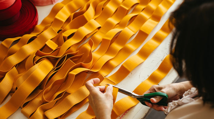 A seamstress uses green scissors to carefully cut a bright ochre length of elastic from long strips laid across a table.